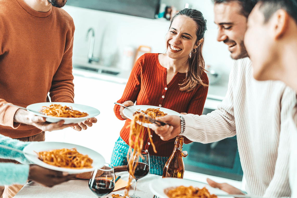 Gruppo di amici sorridenti attorno a una tavola apparecchiata mentre condividono un piatto di pasta e un bicchiere di vino.