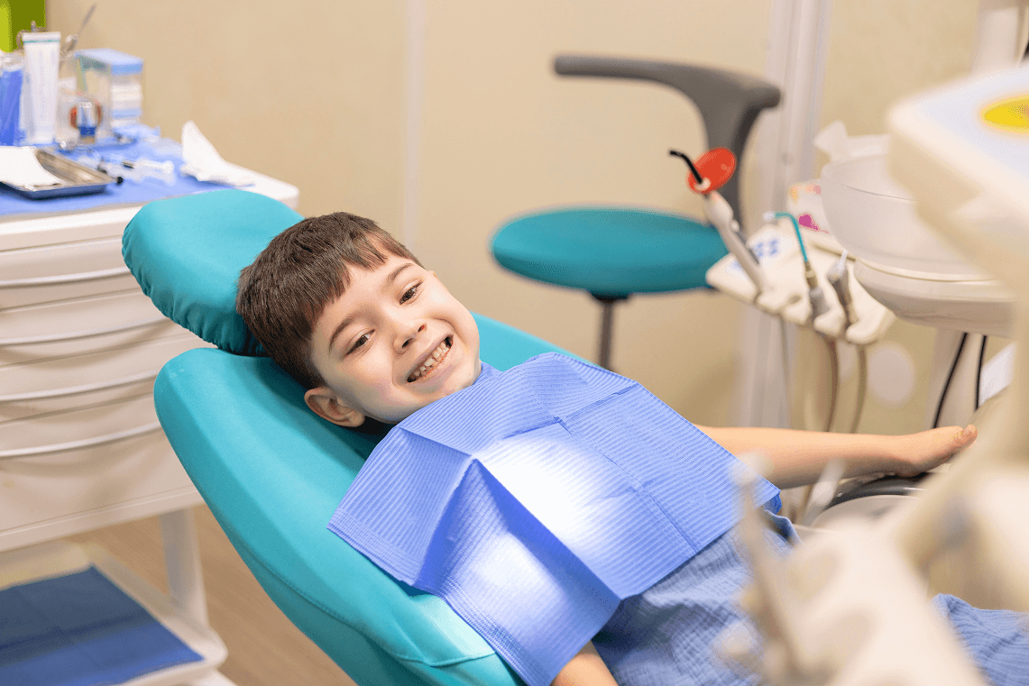 Bambino sorridente e rilassato sulla poltrona da dentista celeste con bavaglino di protezione blu.
