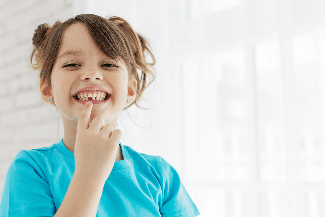 Bambina sorridente in maglia azzurra che indica con il dito la caduta di un dente da latte dell'arcata superiore.
