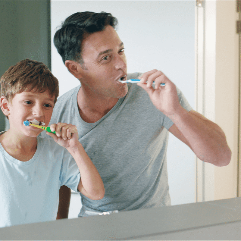 Un padre e il suo giovane figlio che utilizzano spazzolini manuali lavandosi i denti insieme davanti allo specchio del bagno.