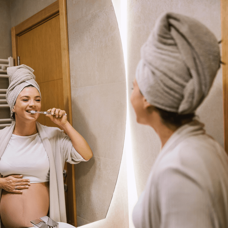 Donna in gravidanza con asciugamano in testa che sorride e si lava i denti davanti allo specchio del bagno di casa.
