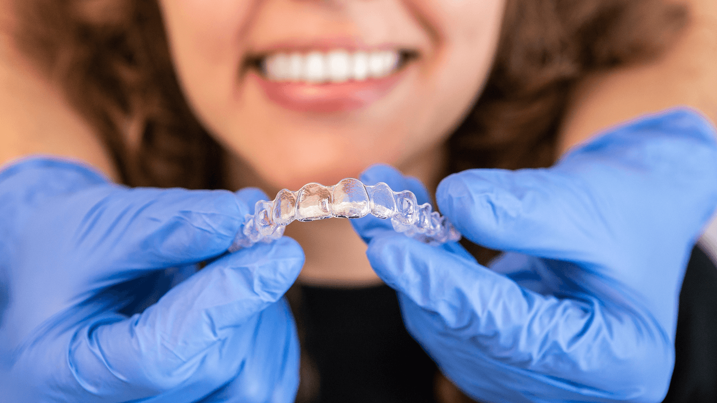 Primo piano delle mani di un dentista con guanti azzurri che tengono una mascherina trasparente per l'ortodonzia invisibile, con il sorriso del paziente sfocato sullo sfondo.