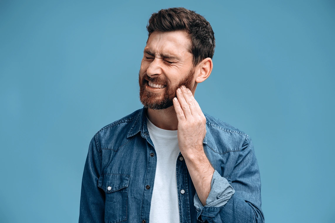 Uomo con la barba e camicia di jeans che prova dolore e si tocca la guancia a causa di un forte mal di denti.