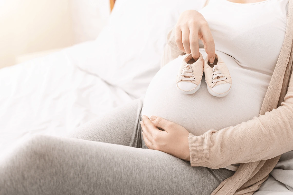 Dettaglio del pancione di una donna in gravidanza che sostiene un paio di scarpine da neonato rosa.