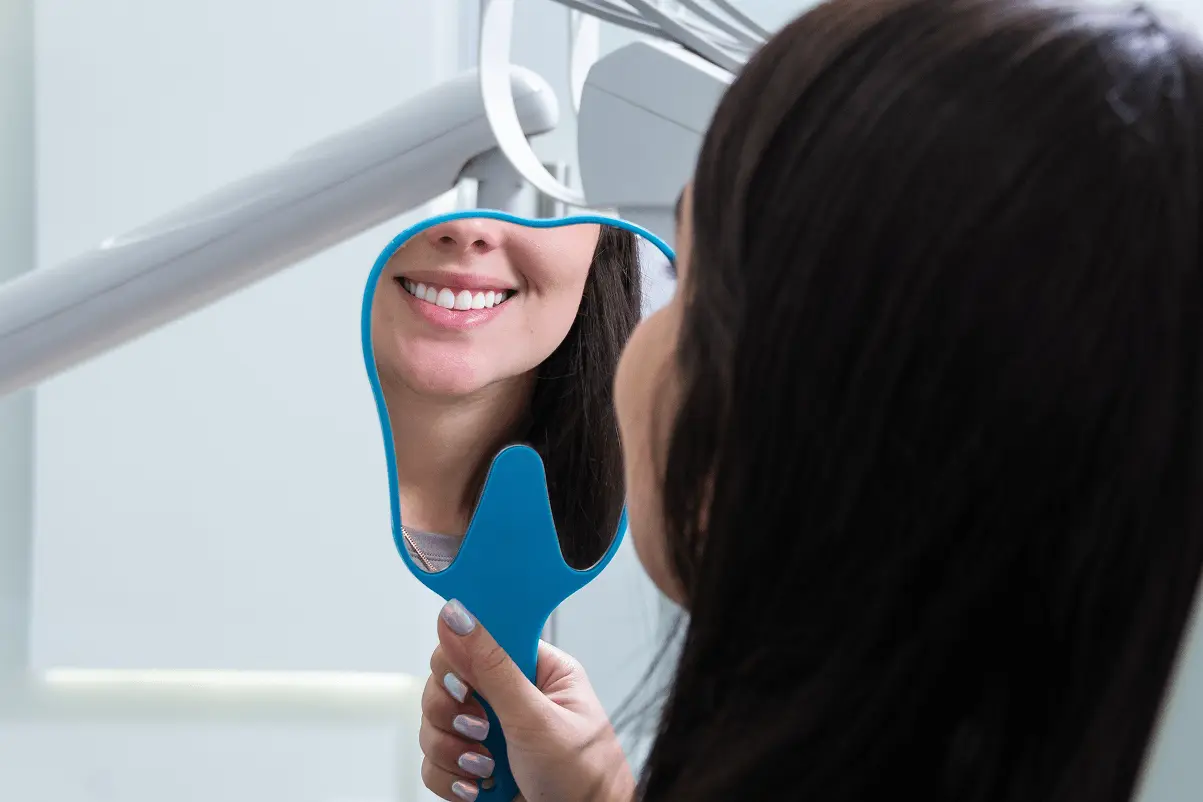 Vista da dietro di una donna che guarda il suo sorriso perfetto e denti bianchi riflessi in uno specchio a mano azzurro a forma di dente.