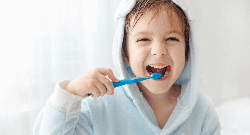 Bambino sorridente con accappatoio celeste con cappuccio che si lava i denti con uno spazzolino blu manuale.