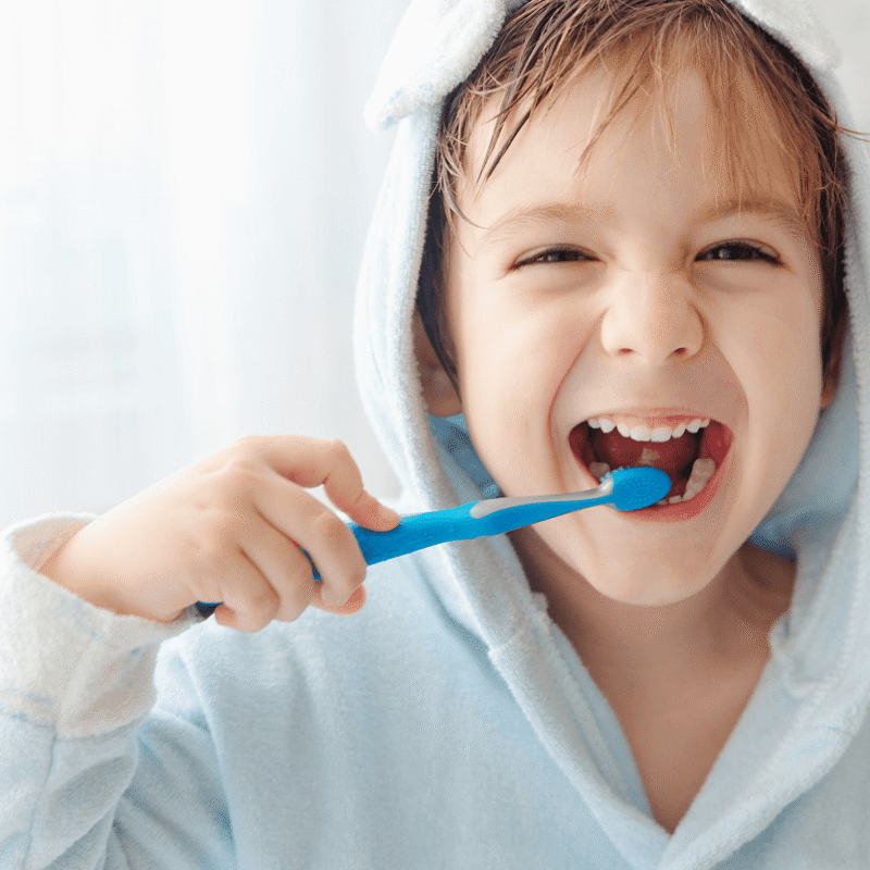 Bambino sorridente con accappatoio celeste con cappuccio che si lava i denti con uno spazzolino blu manuale.