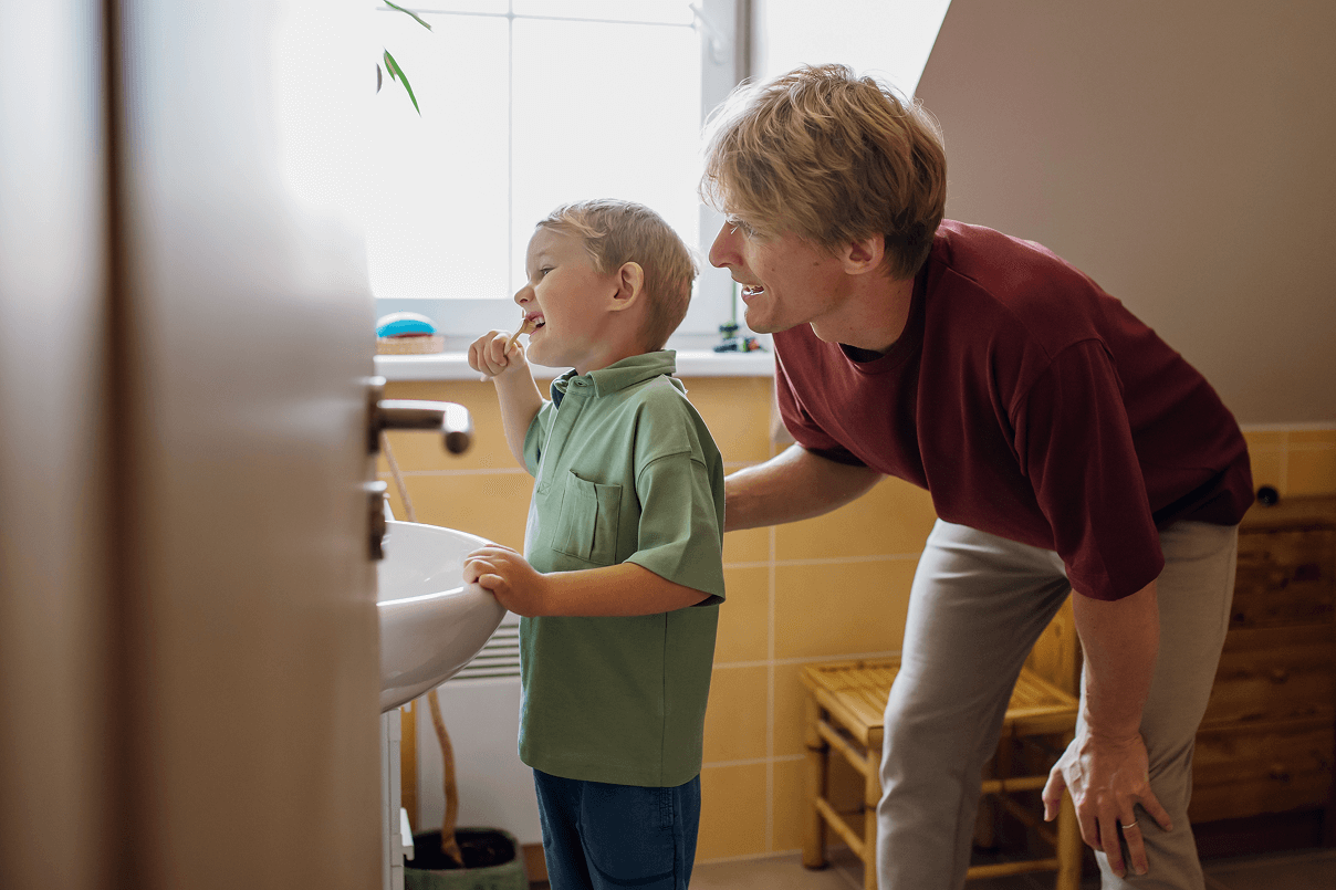 Vista di profilo di un bambino con polo verde che si lava i denti al lavandino del bagno mentre il padre al suo fianco lo guarda e lo incoraggia.