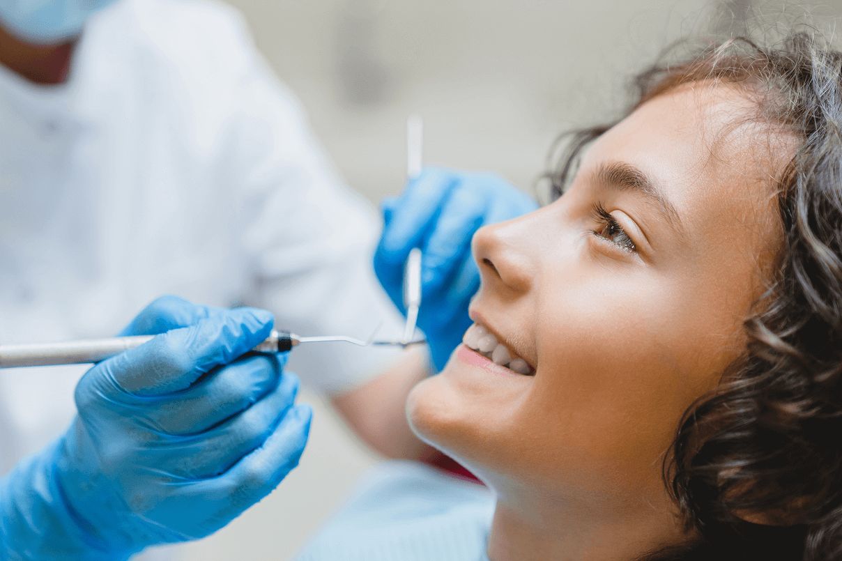 Primo piano di profilo di una giovane paziente sorridente con capelli ricci durante un controllo dei denti effettuato dal dentista con guanti blu e specchietto.