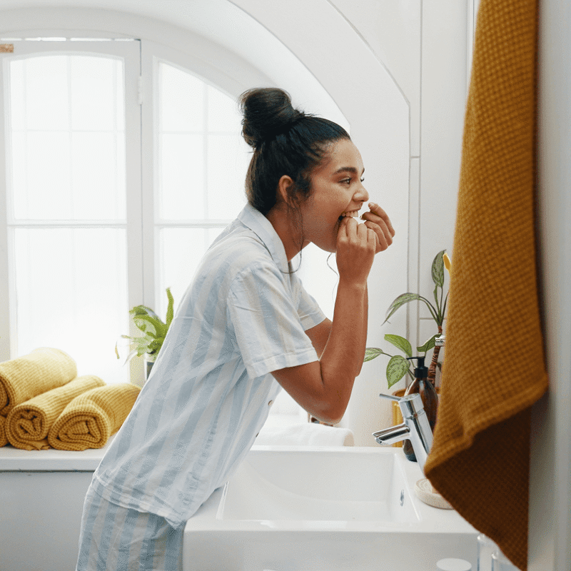 Vista di profilo di una ragazza in pigiama a righe che utilizza il filo interdentale davanti allo specchio del bagno di casa.