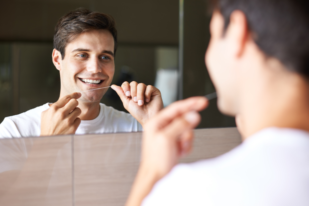 Un giovane uomo sorridente davanti allo specchio del bagno mentre passa il filo interdentale per la pulizia quotidiana dei denti.