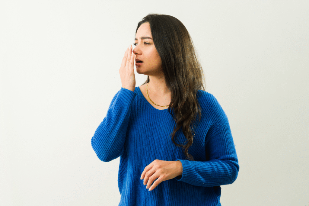 Giovane donna con maglione blu che avvicina la mano alla bocca per controllare la freschezza del proprio alito.