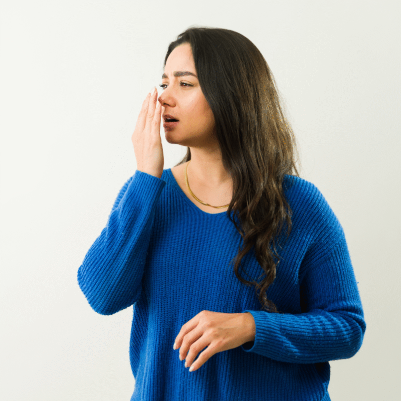 Giovane donna con maglione blu che avvicina la mano alla bocca per controllare la freschezza del proprio alito.