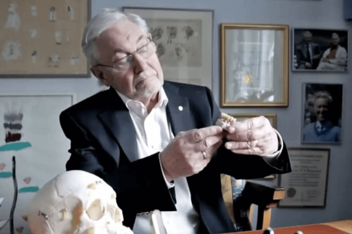 Il Dottor Per-Ingvar Brånemark in giacca e occhiali mentre osserva un modello di protesi dentale e mandibola nel suo studio professionale.