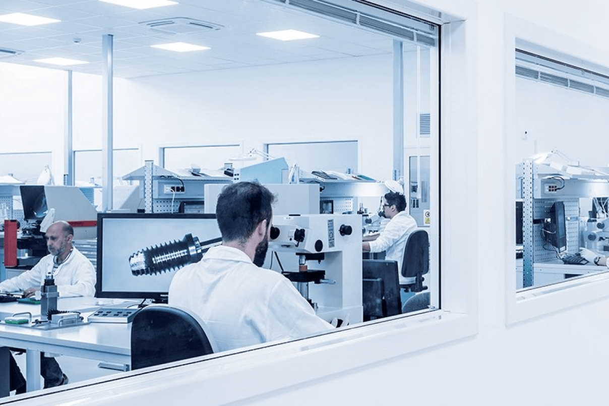 Vista attraverso una vetrata di operatori in camice bianco al lavoro in un laboratorio sterile con microscopi e monitor con la scansione di un impianto dentale.