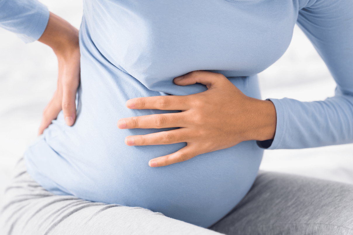 Dettaglio del pancione di una donna in gravidanza vestita di azzurro mentre si tiene una mano sulla pancia e l'altra sulla zona lombare per il mal di schiena.
