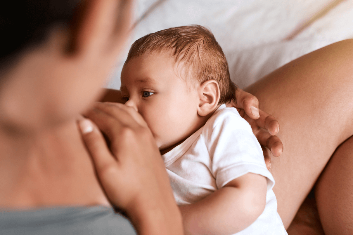 Vista da sopra le spalle della madre mentre tiene in braccio e allatta al seno il suo neonato con un body bianco.