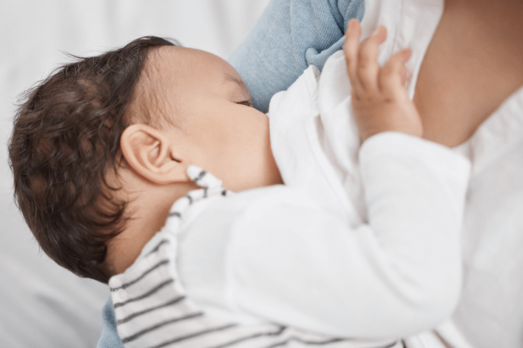 Primo piano di un neonato con maglia a righe e maniche bianche mentre viene allattato al seno tra le braccia della madre.