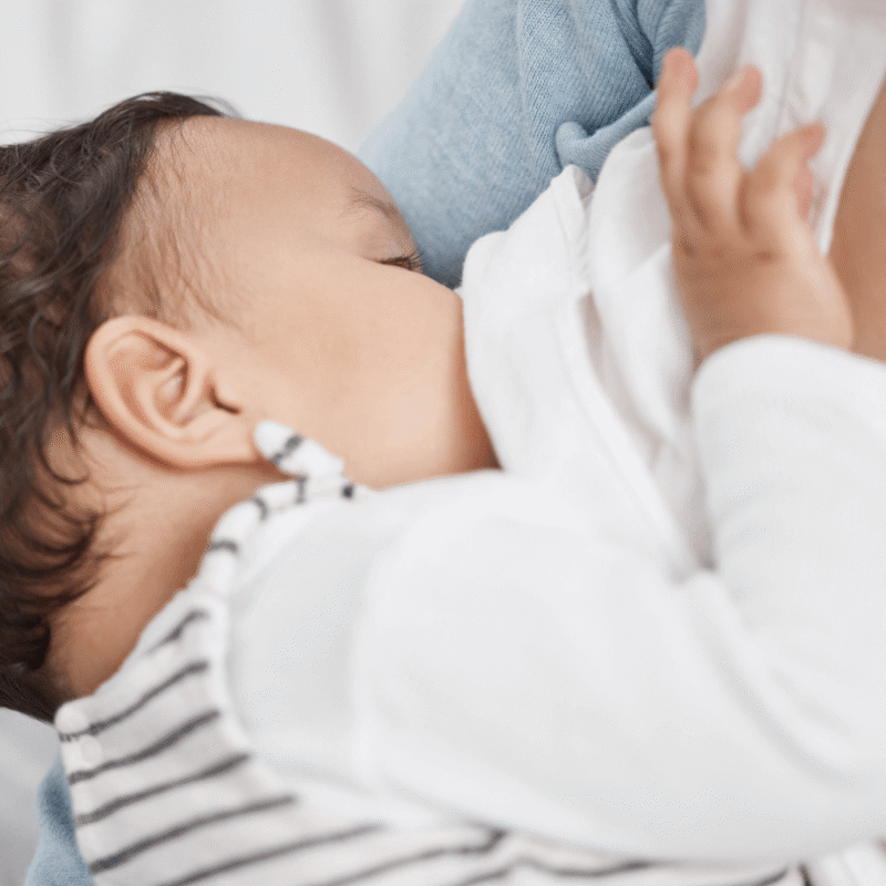 Primo piano di un neonato con maglia a righe e maniche bianche mentre viene allattato al seno tra le braccia della madre.