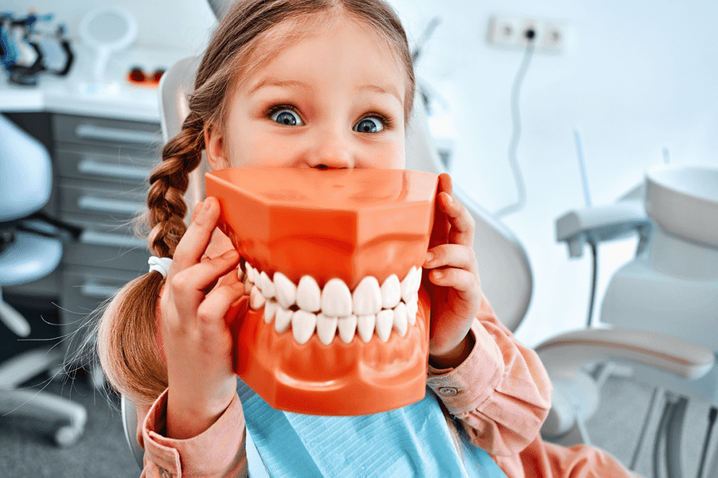 Bambina con le trecce seduta sulla poltrona del dentista che gioca nascondendosi il viso dietro un grande modello arancione di dentiera.