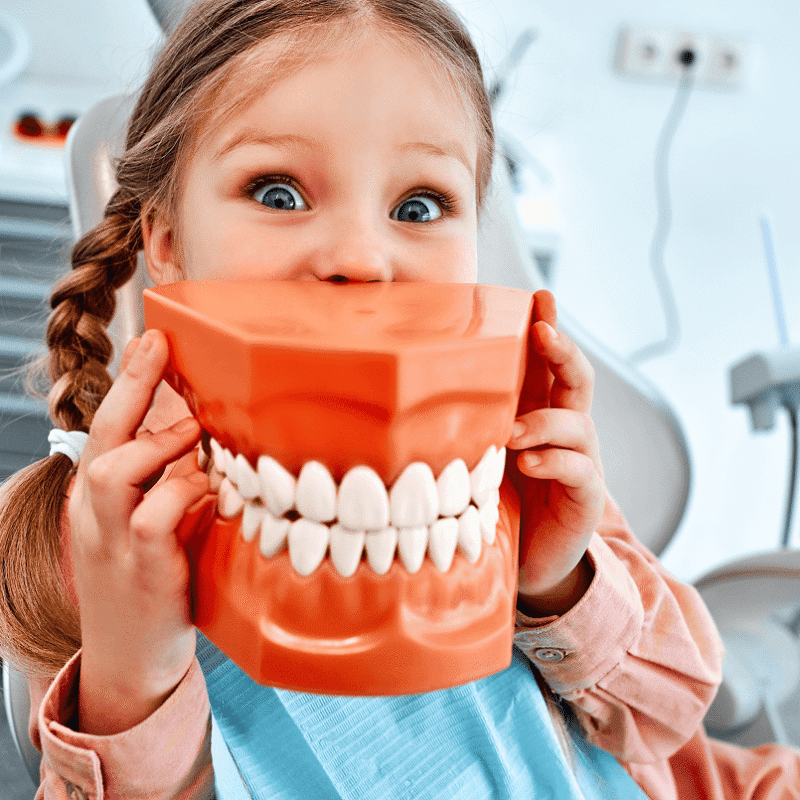 Bambina con le trecce seduta sulla poltrona del dentista che gioca nascondendosi il viso dietro un grande modello arancione di dentiera.