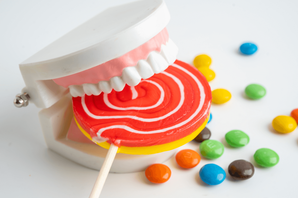 Modello anatomico di dentiera plastica che morde un lecca-lecca gigante colorato circondato da confetti di zucchero multicolor.