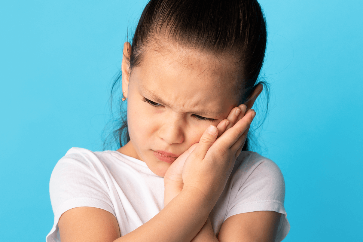Bambina con t-shirt bianca che si tiene le mani sulla guancia con un'espressione sofferente a causa del mal di denti, su sfondo azzurro.