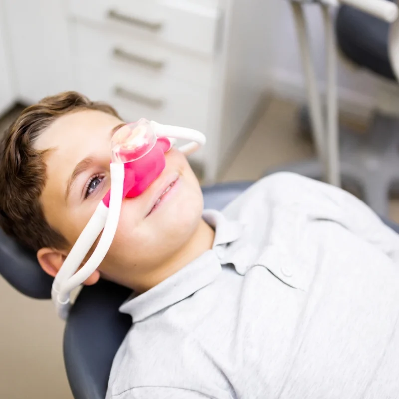 Bambino sorridente disteso sulla poltrona da dentista mentre indossa una mascherina nasale rosa per la sedazione cosciente.