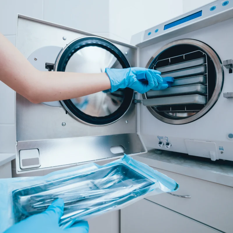 Assistente con guanti azzurri preleva vasi di strumenti dall'autoclave tenendo in mano una busta sterile sigillata.