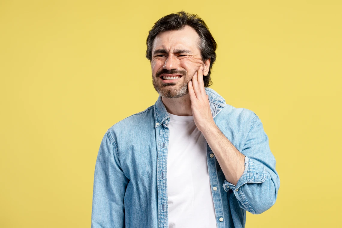 Uomo con camicia in di jeans che prova un forte mal di denti e si tiene la mano sulla guancia con un'espressione dolorante su sfondo giallo.