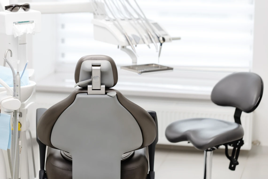 Vista posteriore di una poltrona da visita odontoiatrica moderna in pelle grigia con sgabello per il medico e manipoli dentistici sullo sfondo.